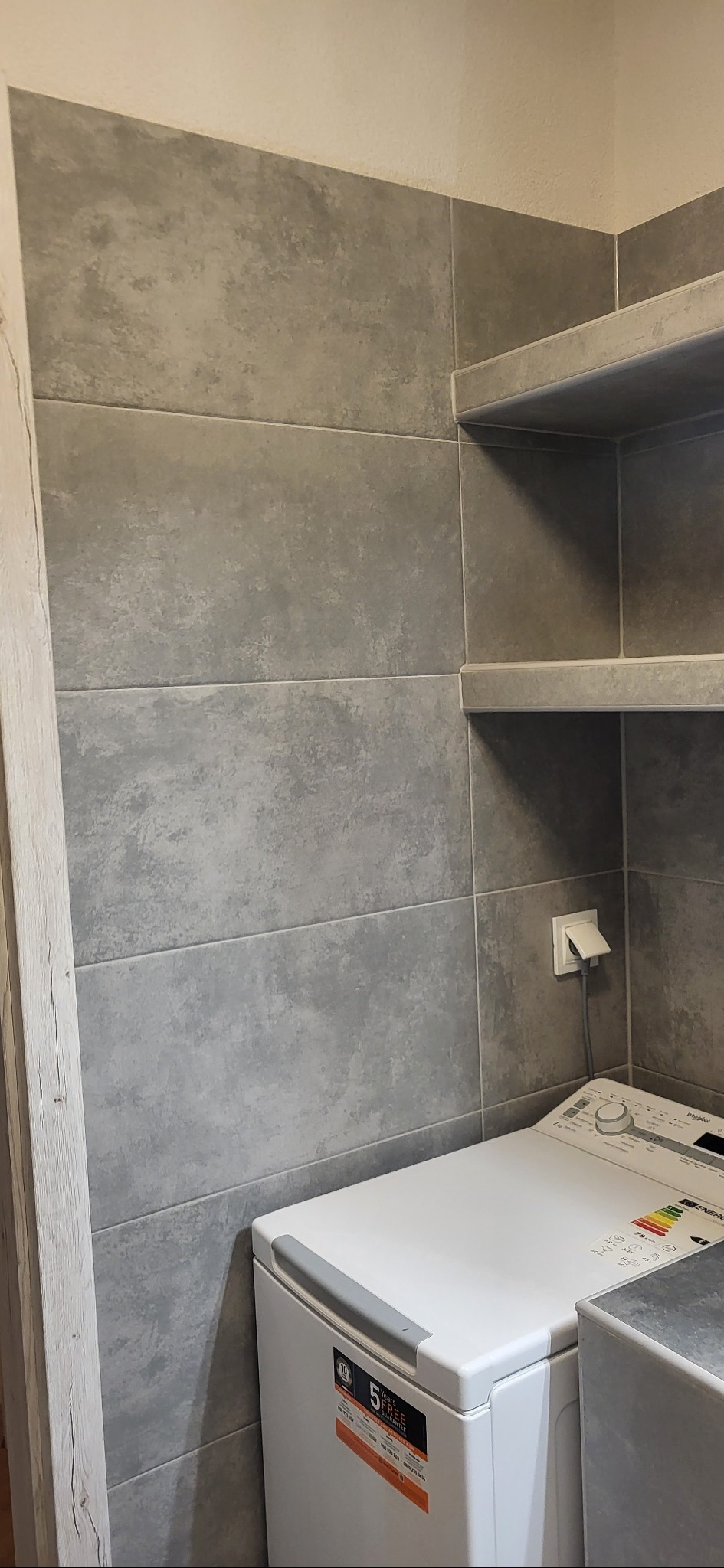 Szare, poziome płytki w łazience z wnękami na półki. Widoczna pralka i gniazdko elektryczne. Minimalistyczny design, neutralna kolorystyka.