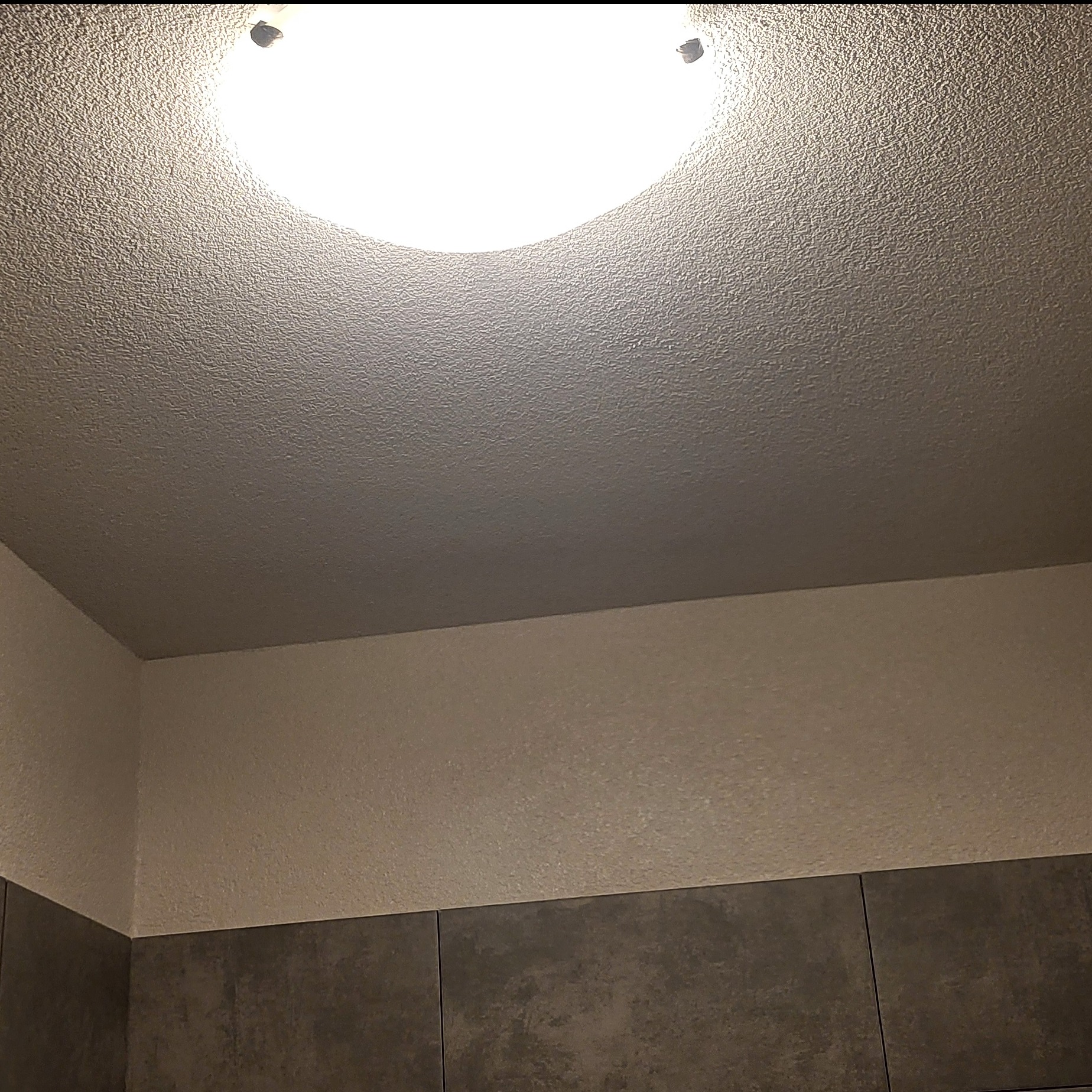 Oświetlony sufit z fakturą baranka i fragment ściany z szarymi płytkami w dolnej części. Widok z dołu, wnętrze po remoncie.