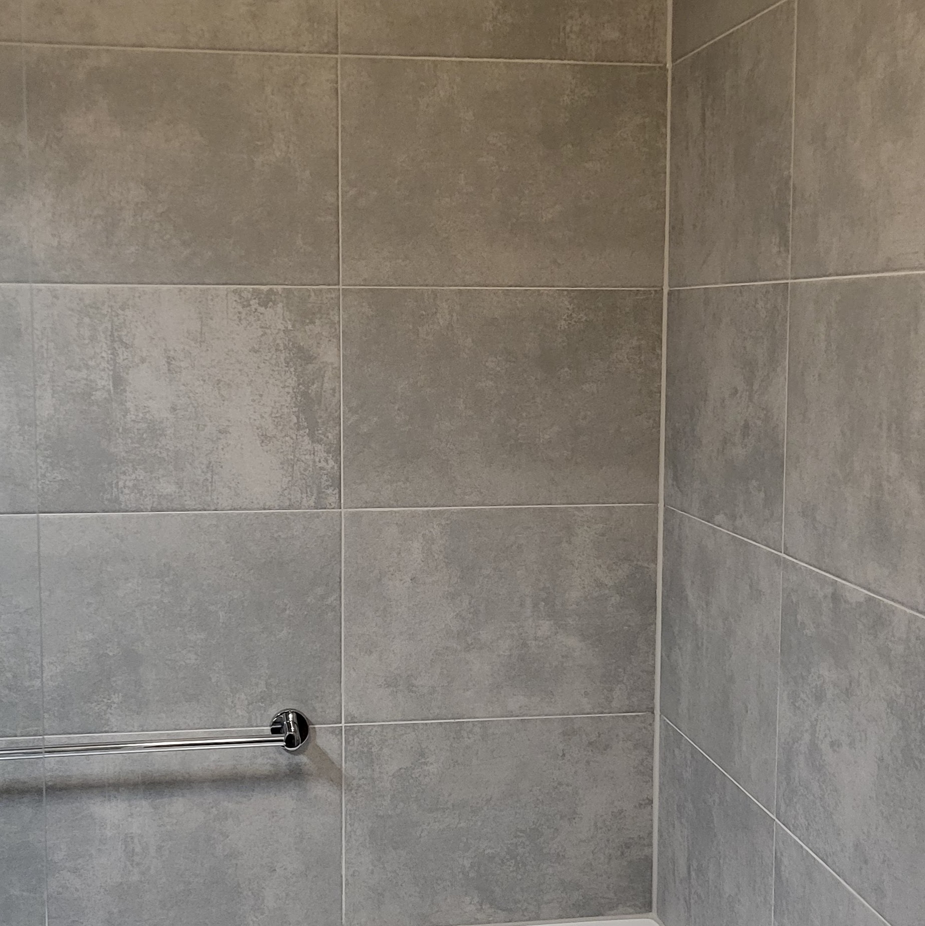 Szare, matowe płytki w łazience z chromowanym uchwytem na ręczniki. Minimalistyczny design, fuga w kolorze zbliżonym do płytek. Ujęcie z narożnika.