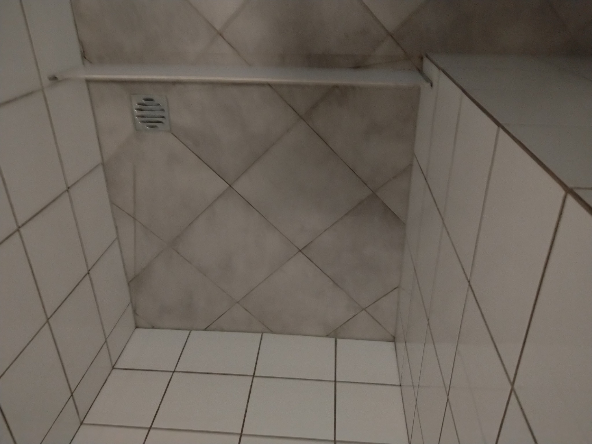 Wykończona kabina prysznicowa z jasnymi, kwadratowymi płytkami na ścianach i podłodze, ułożonymi prosto i romboidalnie. Widoczny odpływ liniowy.