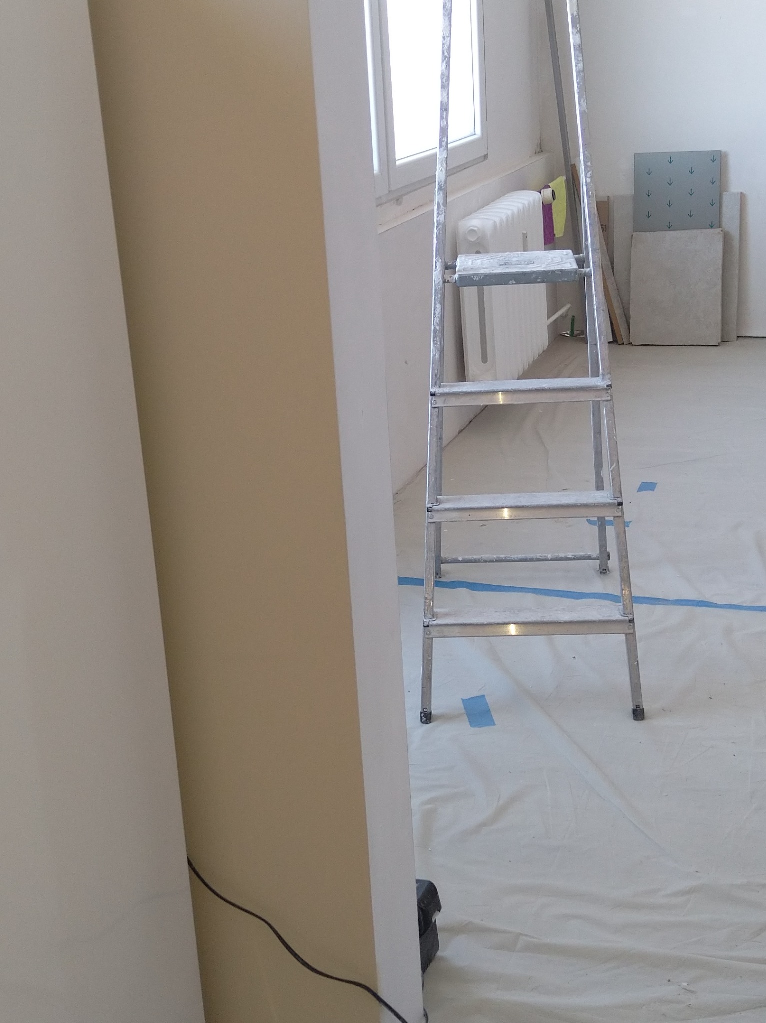 Przygotowanie do malowania: metalowa drabina stoi na folii ochronnej, widoczne płytki i grzejnik. Ściana w trakcie malowania, częściowo w jasnym beżu.