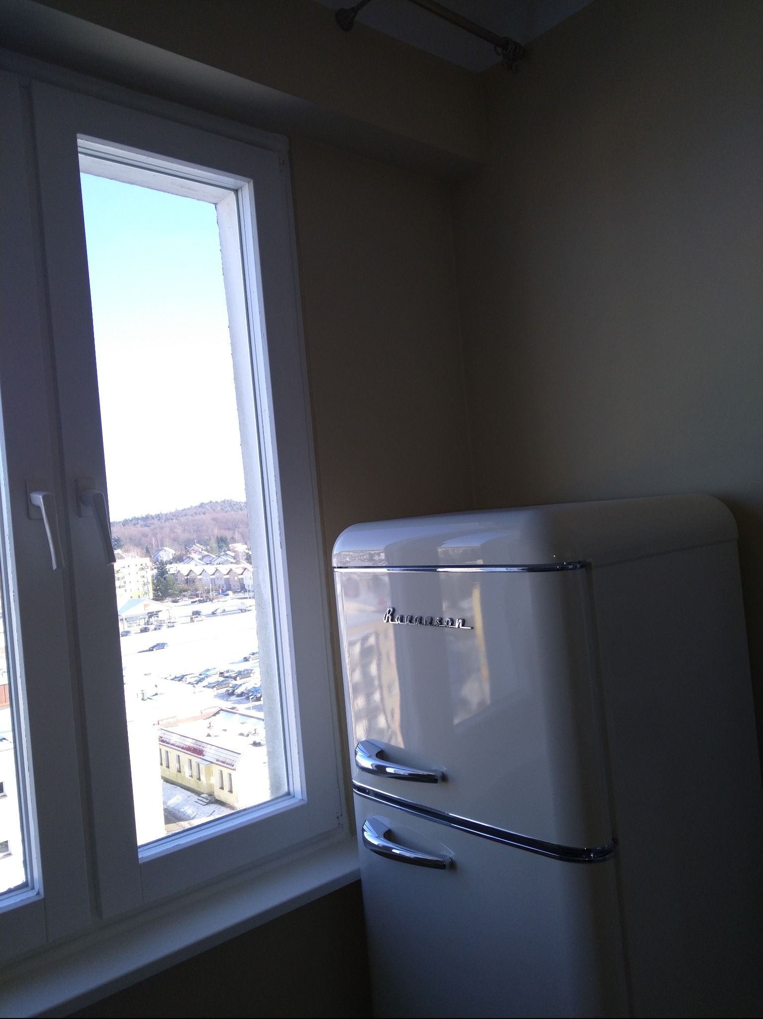 Retro lodówka marki Ravanson w jasnym wnętrzu, widoczna z boku przy oknie z widokiem na miasto. Charakterystyczne chromowane uchwyty i zaokrąglone kształty.