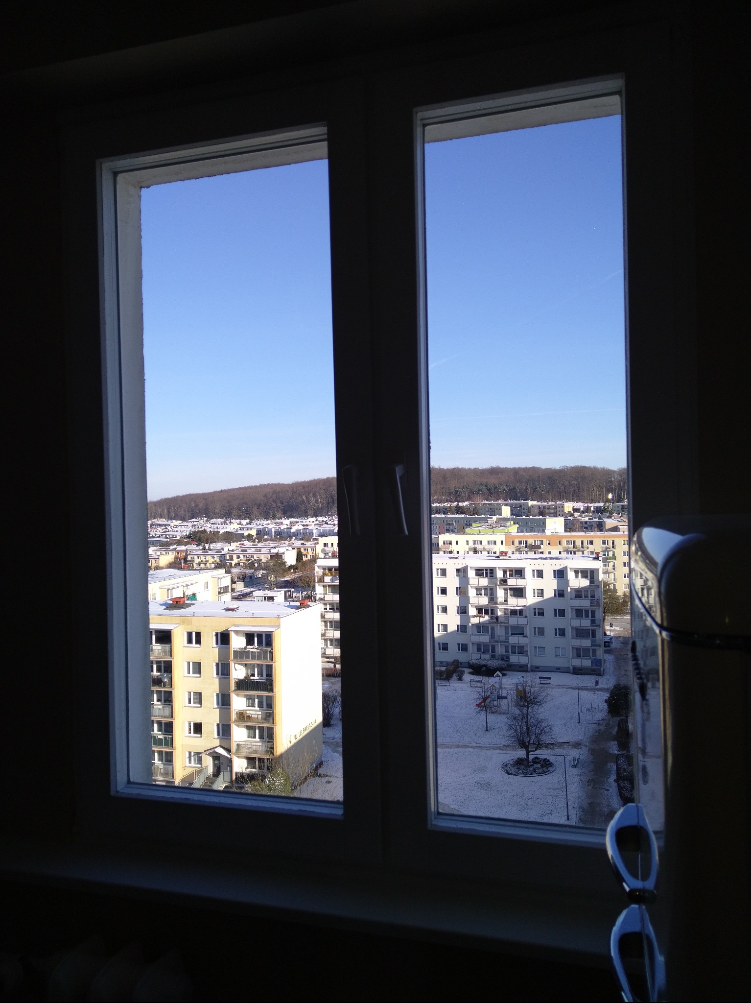 Widok z okna na osiedle w zimowej scenerii. Białe budynki i drzewa pokryte śniegiem, błękitne niebo. Okno dwuskrzydłowe, białe ramy. W tle las na wzgórzu.