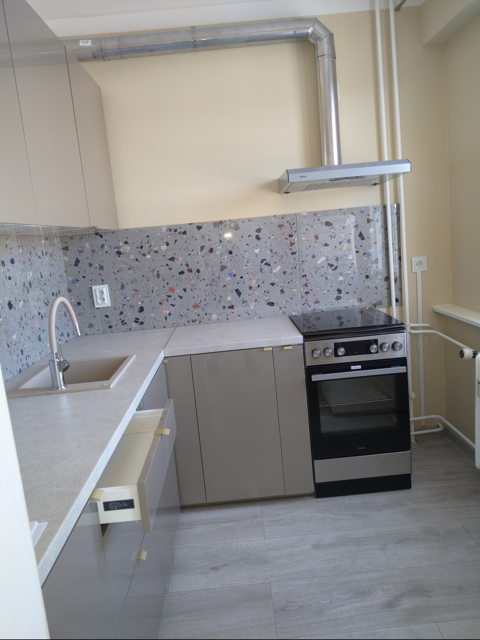 Nowoczesna kuchnia w beżach i szarościach: błyszczące fronty szafek, blat z konglomeratu, mozaika na ścianie i srebrny okap. Otwarta szuflada z widocznym logo.