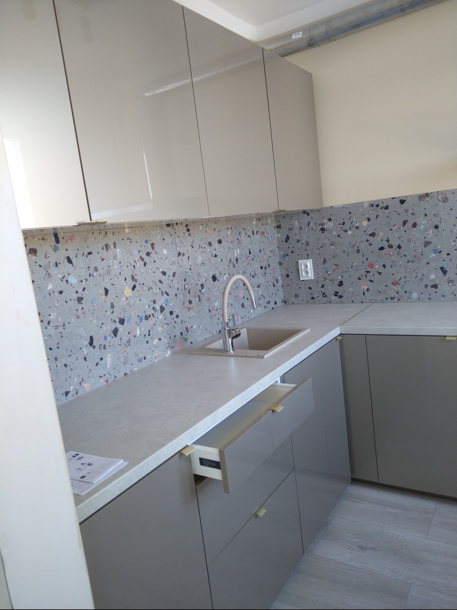 Nowoczesna kuchnia z szarymi, lakierowanymi szafkami, blatem i oryginalną mozaiką na ścianie. Otwarta szuflada z widocznym systemem Blum. Minimalistyczny design.