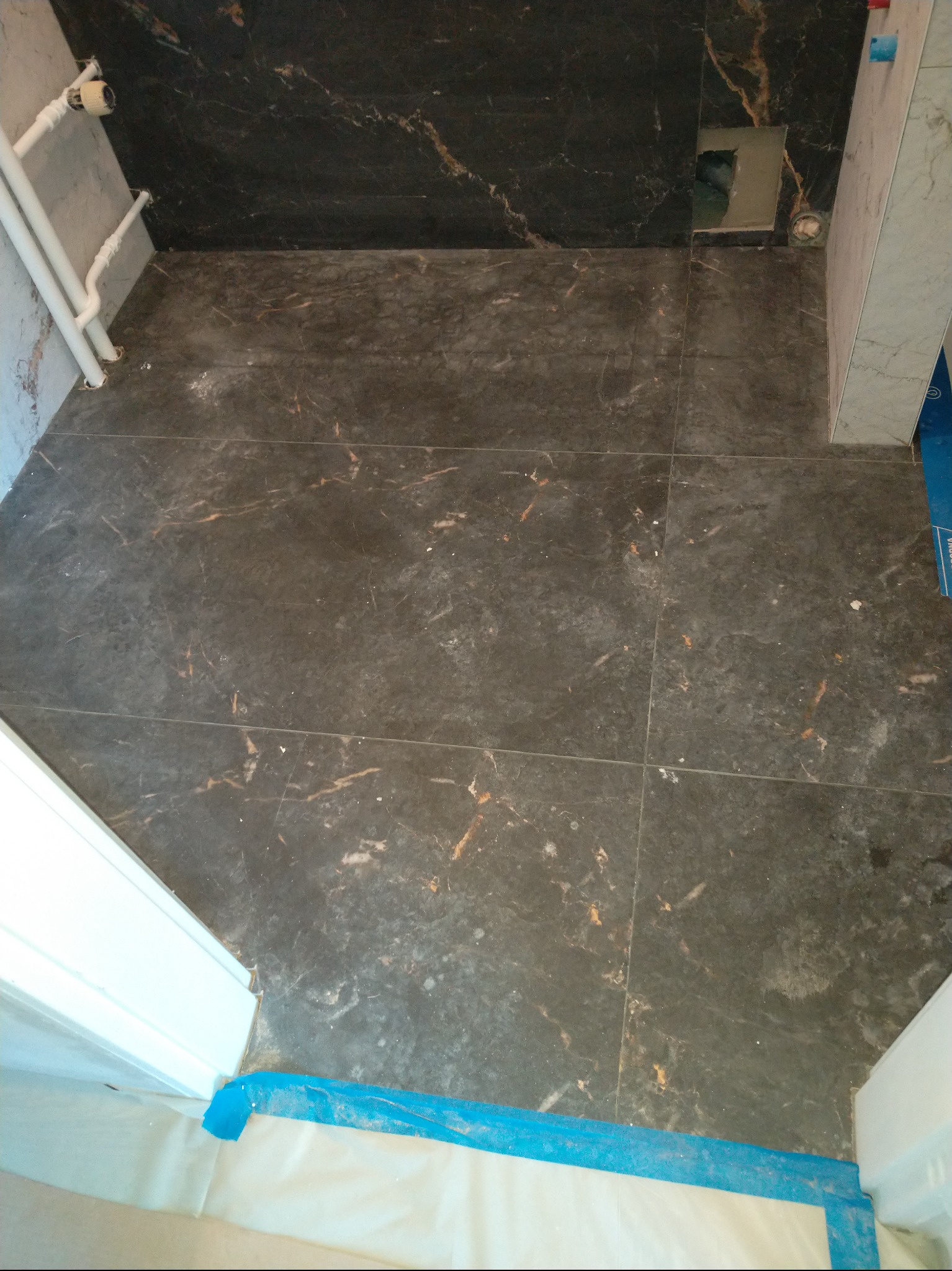 Ułożone, ciemne płytki imitujące marmur na podłodze i ścianie w łazience. Widoczne rury i zabezpieczenia podłogi folią malarską. Górny kadr.