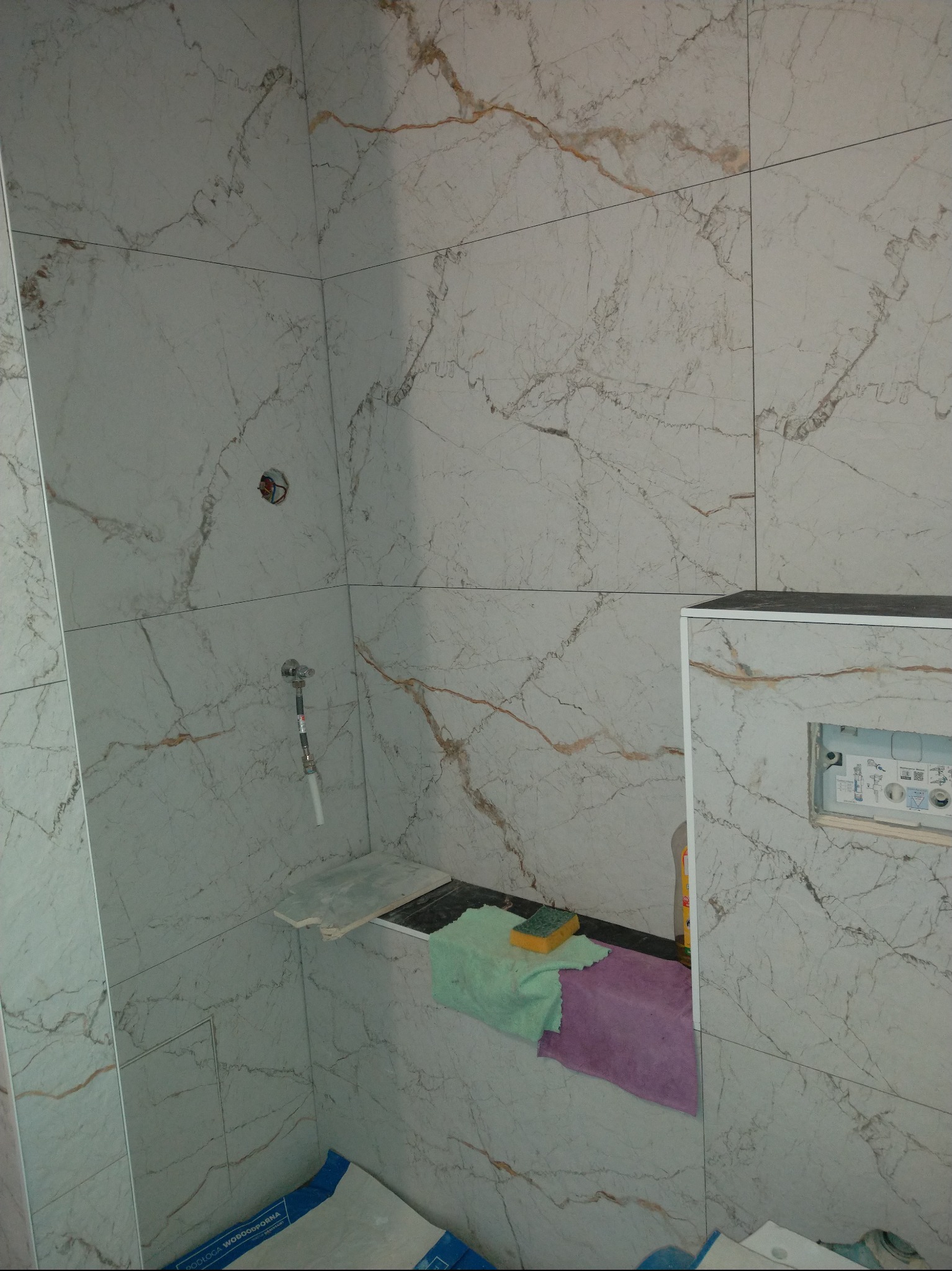 Wykończona kabina prysznicowa z marmurowymi płytkami, widoczne elementy armatury i półka z akcesoriami. Minimalistyczny design z subtelnym wzorem żyłkowania.