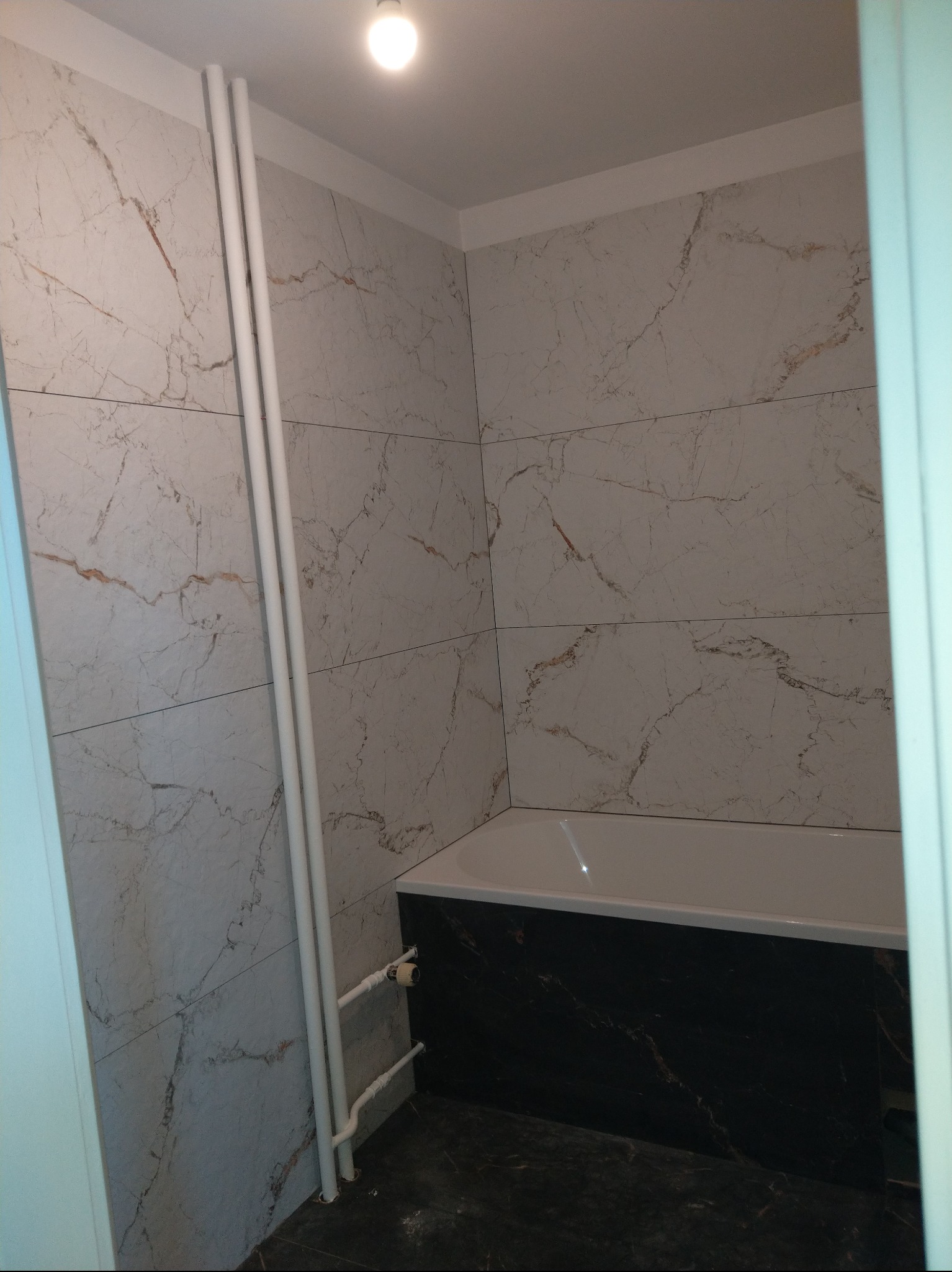 Wykończona łazienka z wanną obudowaną czarnym marmurem i ścianami wyłożonymi jasnymi płytkami imitującymi marmur. Widoczne białe rury grzewcze.