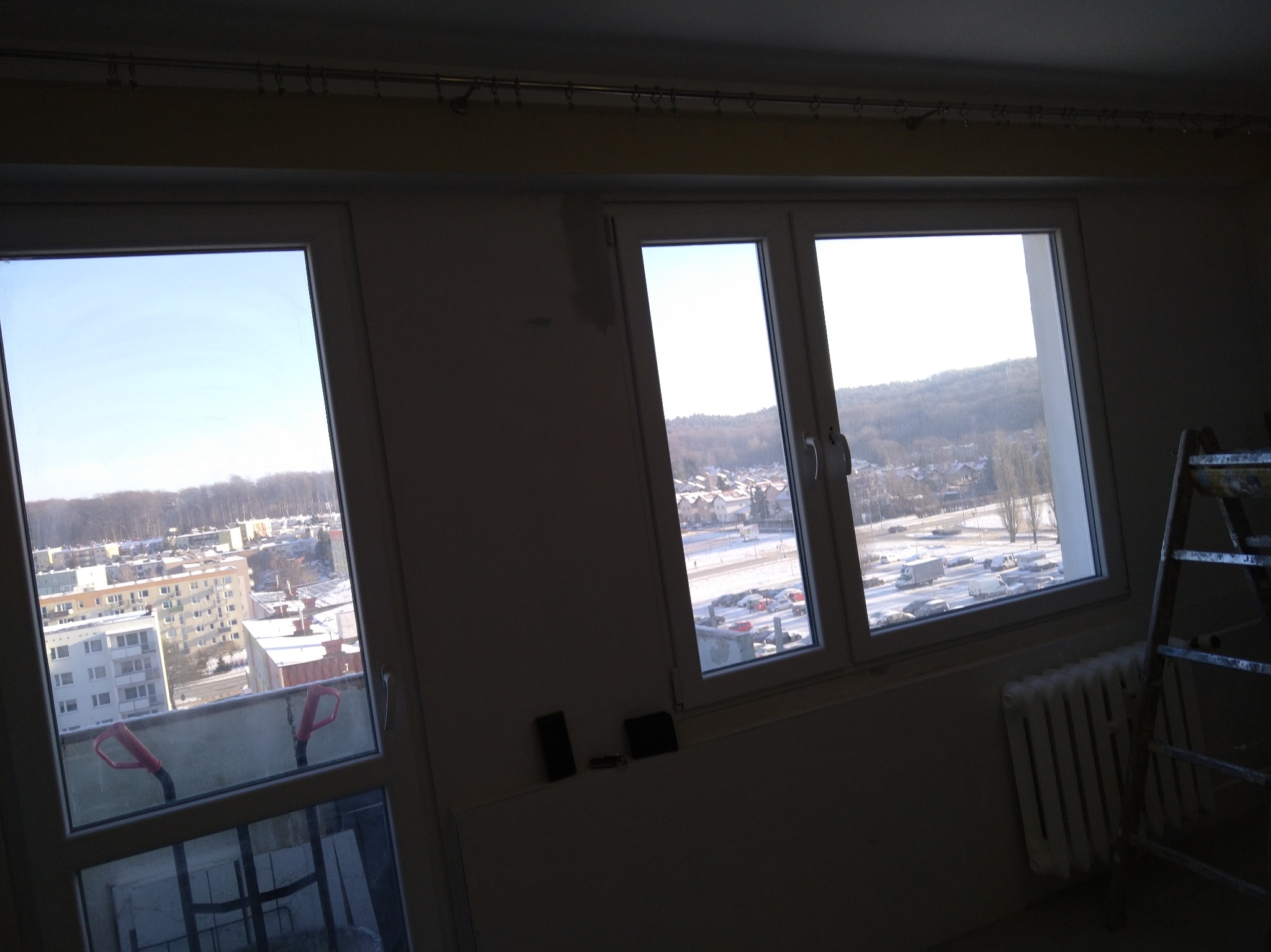 Wnętrze w trakcie remontu w Gąskach: dwa okna z widokiem na miasto, grzejnik i drabina malarska. Ściany przygotowane do malowania.