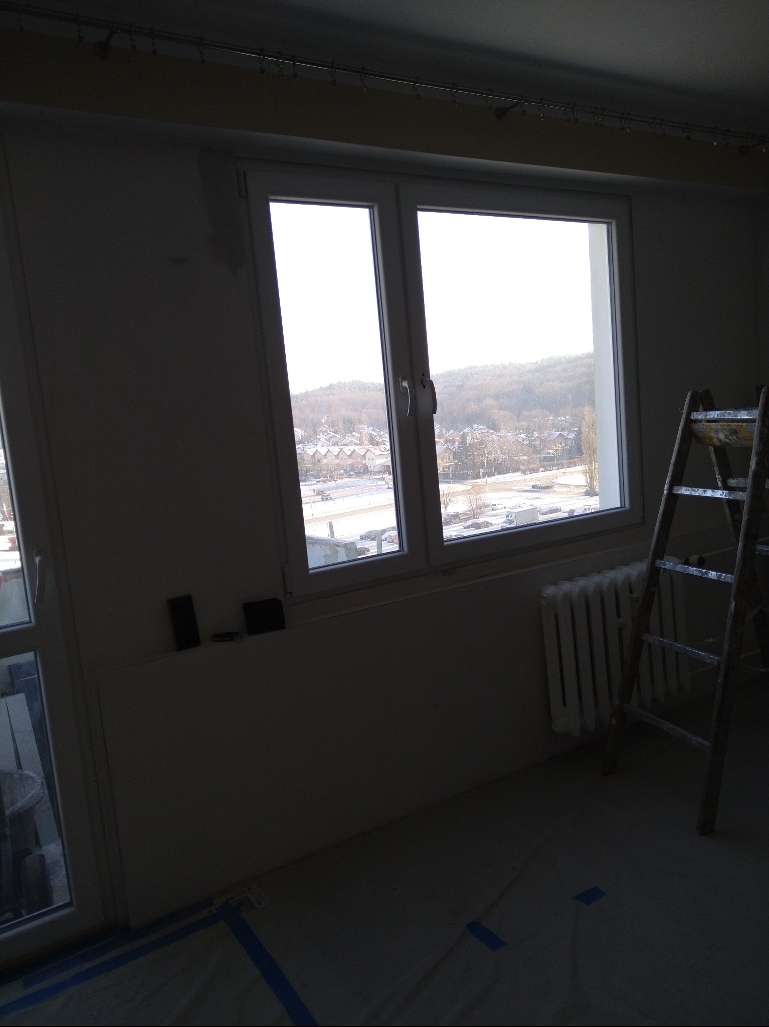 Pokój w trakcie prac. Okno z widokiem na miasto, drabina oparta o ścianę, zabezpieczona podłoga. Przygotowania do malowania lub wykończenia.