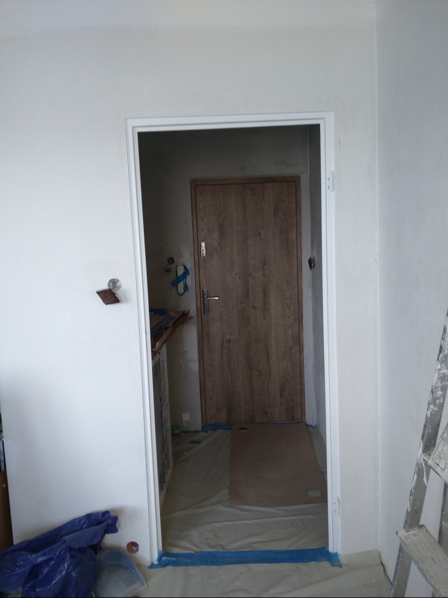 Wnętrze w trakcie prac wykończeniowych: zamontowane ościeżnice drzwiowe, zabezpieczona podłoga folią malarską, ściany przygotowane do malowania. Widoczna drabina malarska.