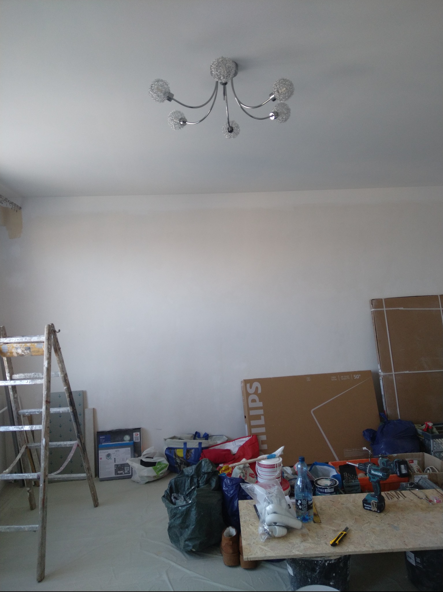 Biała ściana po malowaniu z widoczną drabiną, narzędziami i kartonami. Nowoczesna lampa sufitowa. Przygotowania do dalszych prac wykończeniowych.