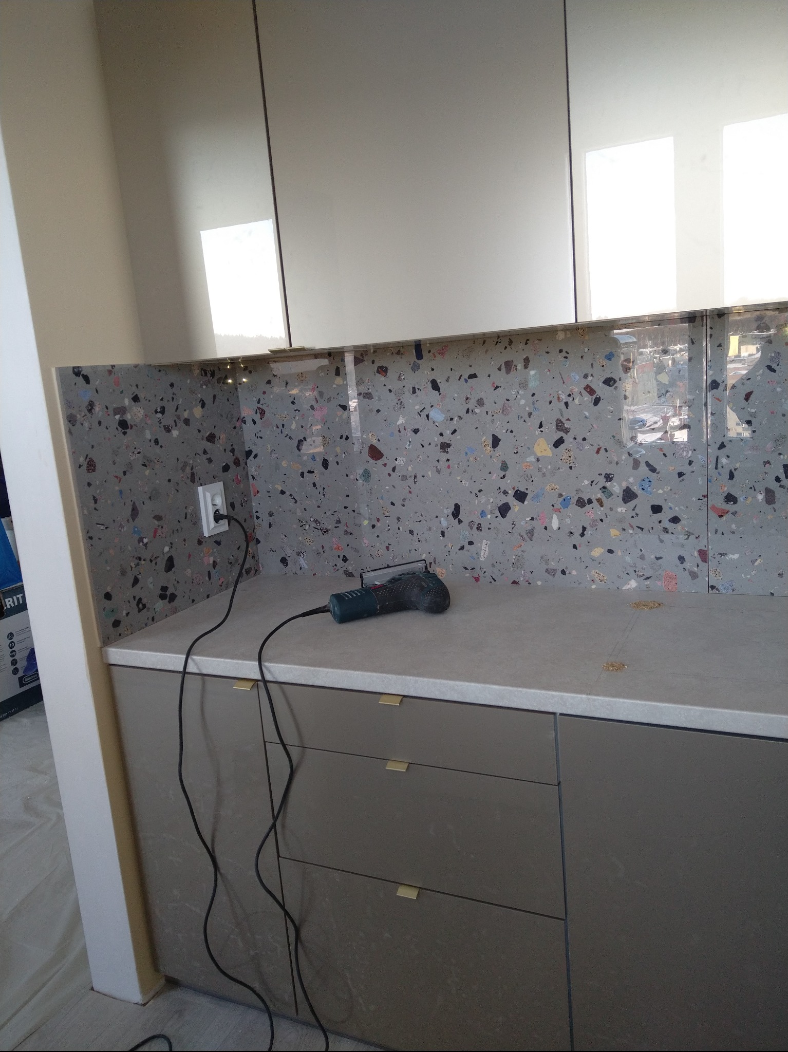 Kuchnia w trakcie wykańczania: szara mozaika na ścianie nad blatem, szafki w odcieniach szarości i narzędzie do cięcia płytek na blacie. Widoczne gniazdko elektryczne.