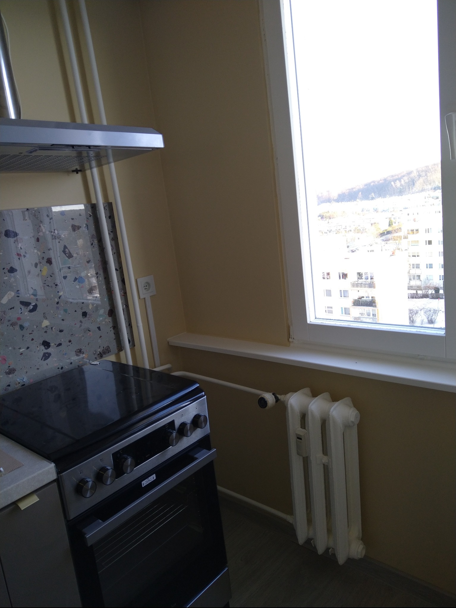 Jasna kuchnia z widokiem na miasto przez okno. Nowoczesna płyta grzewcza i okap nad nią. Biały grzejnik pod oknem. Ściany w odcieniach beżu. Minimalistyczny design.