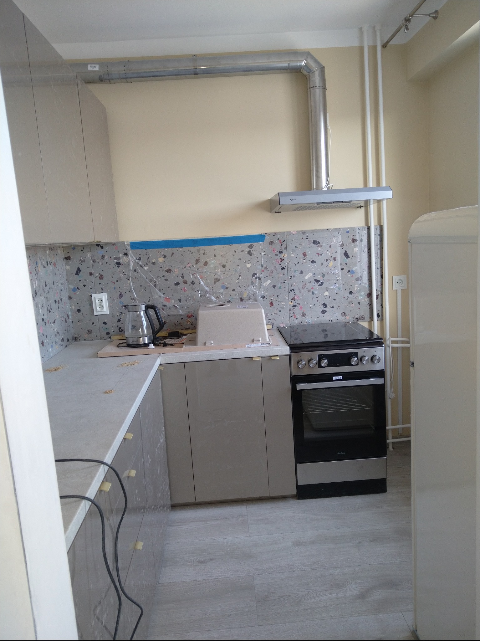 Jasna kuchnia po remoncie: szare szafki z złotymi uchwytami, mozaika na ścianie, srebrny okap i płyta grzewcza. Widoczne drobne ślady po remoncie.