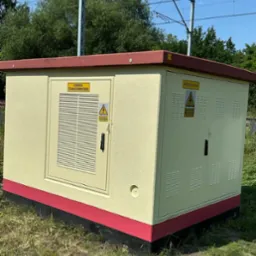 Pomalowana na beżowo komora transformatorowa z czerwonymi akcentami, otoczona zielenią. Widoczne napisy ostrzegawcze i elementy infrastruktury kolejowej w tle.