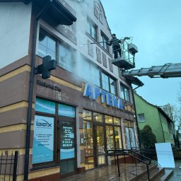 Horizon Build Tomasz Zdanowicz - Czyszczenie elewacji apteki z podnośnika koszowego. Pracownik spryskuje ścianę wodą pod ciśnieniem, widoczne pojemniki z płynami i mokre schody.