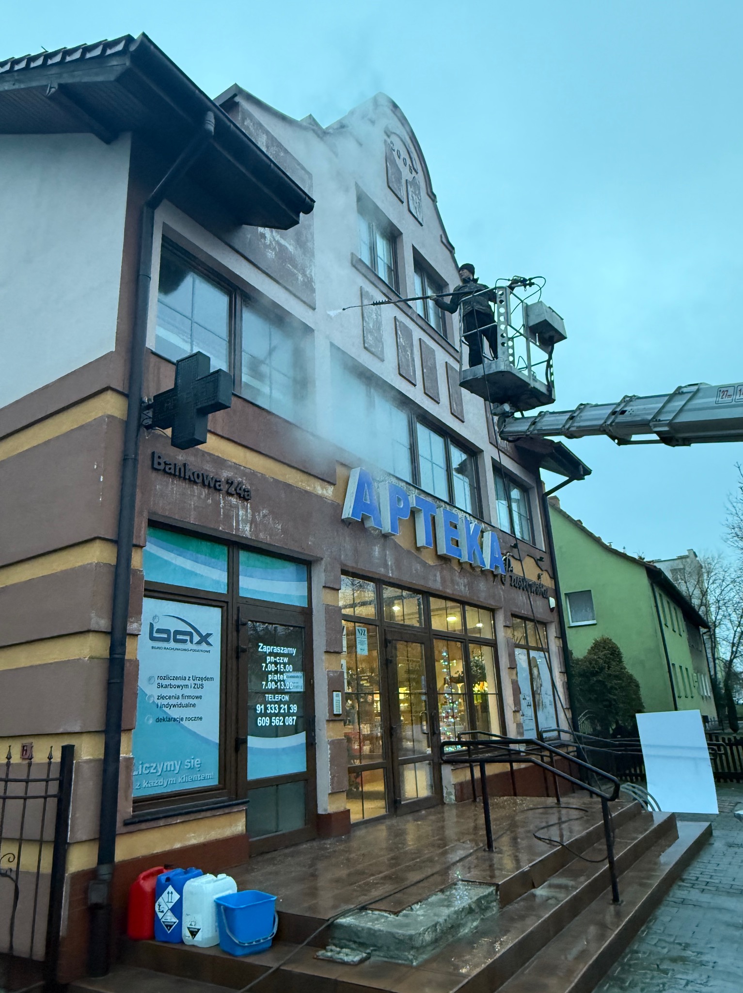 Czyszczenie elewacji apteki z podnośnika koszowego. Pracownik spryskuje ścianę wodą pod ciśnieniem, widoczne pojemniki z płynami i mokre schody.