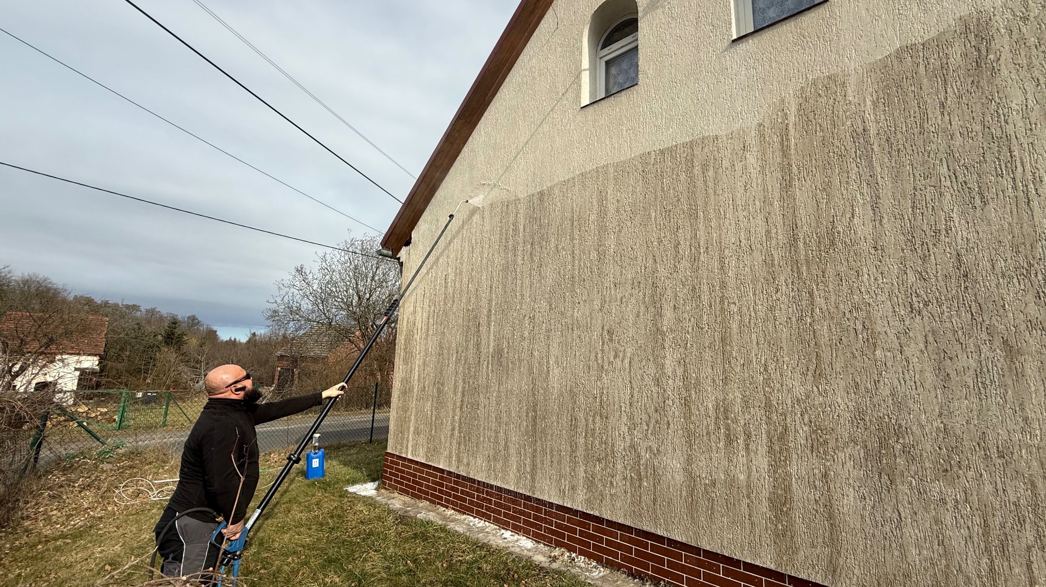 Mężczyzna w okularach myje elewację domu za pomocą lancy teleskopowej. Widoczny efekt czyszczenia: górna część ściany jaśniejsza od dolnej, ceglana podmurówka.