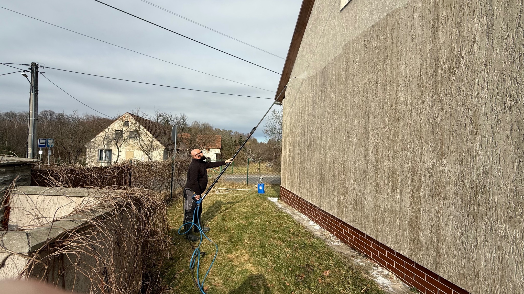 Mężczyzna czyści elewację domu za pomocą lancy ciśnieniowej. Widoczny budynek w tle i niebieski pojemnik na detergent. Praca wykonywana z poziomu trawnika.