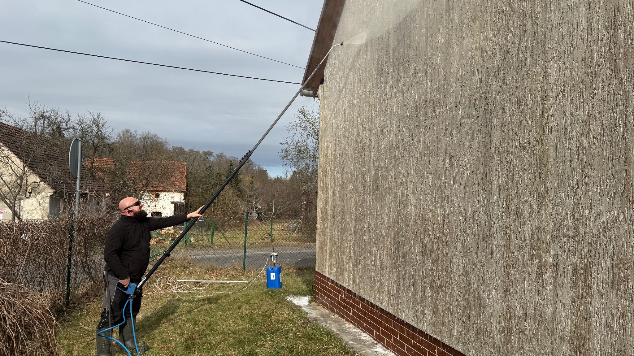 Mężczyzna czyści elewację domu za pomocą lancy ciśnieniowej. Widoczny efekt mycia na teksturowanej ścianie, zarys ceglanej podmurówki i sprzęt do mycia.