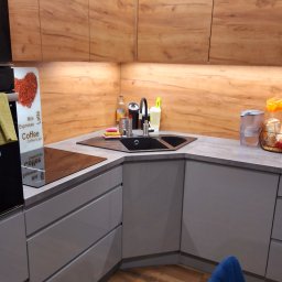 Pure & Fresh Anna Sós - Nowoczesna kuchnia w odcieniach szarości i drewna. Uwagę przykuwa czarny zlew narożny i oświetlenie LED pod szafkami, tworzące ciepłą atmosferę. Widoczny fragment krzesła.