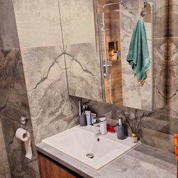 Pure & Fresh Anna Sós - Umywalka z baterią i akcesoriami łazienkowymi na blacie z szarą fakturą, widok na lustro z odbiciem kabiny prysznicowej i ręcznikiem. Marmurowe płytki na ścianach.