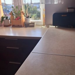 Pure & Fresh Anna Sós - Jasna kuchnia z roślinami w oknie i drewnianym chlebakiem. Blat z delikatnym wzorem, ciemne szafki. Dzień, naturalne światło, firanka z koronką.