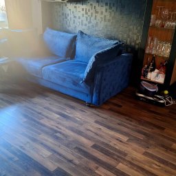 Pure & Fresh - Salon z drewnianą podłogą, niebieską sofą i dekoracyjną ścianą. Na półkach ozdoby i szkło. Świeże, jasne wnętrze po sprzątaniu, gotowe do relaksu.