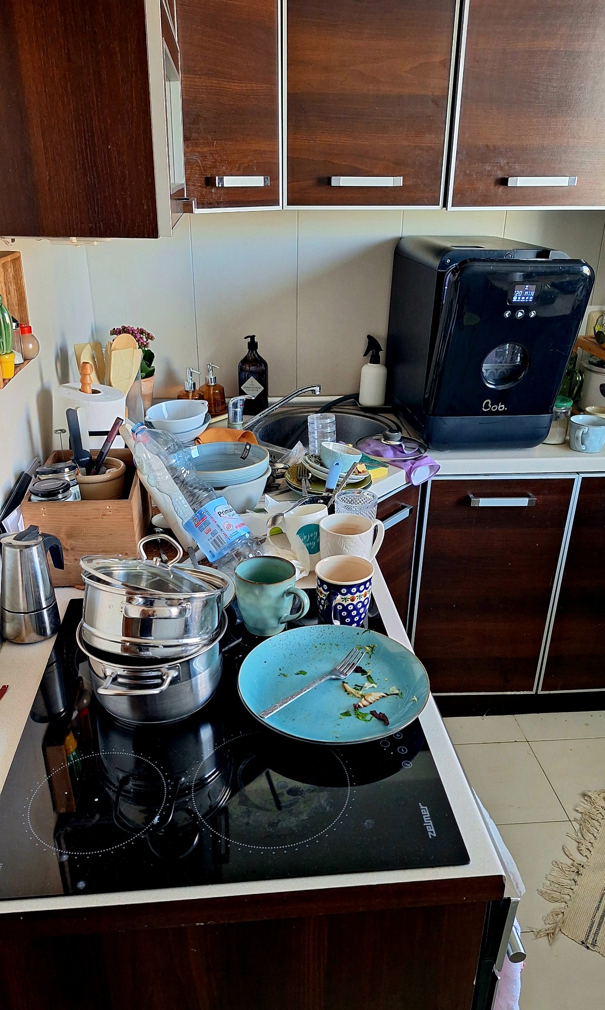 Zastawiony blat kuchenny z naczyniami, garnkami na płycie indukcyjnej i małą zmywarką na blacie. Widoczny bałagan przed sprzątaniem.