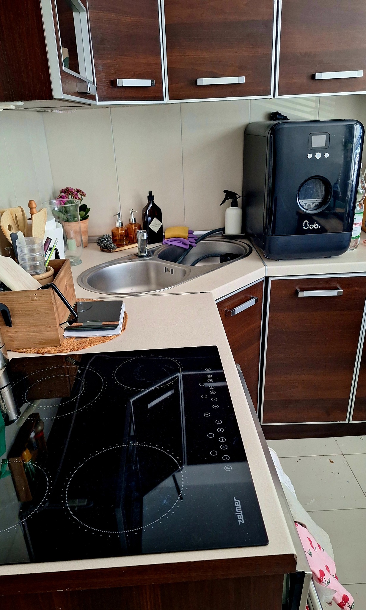 Umyta kuchnia z czarną płytą indukcyjną Zelmer, zlewozmywakiem i małą zmywarką Bob. na blacie. Widoczne drewniane szafki i akcesoria kuchenne.