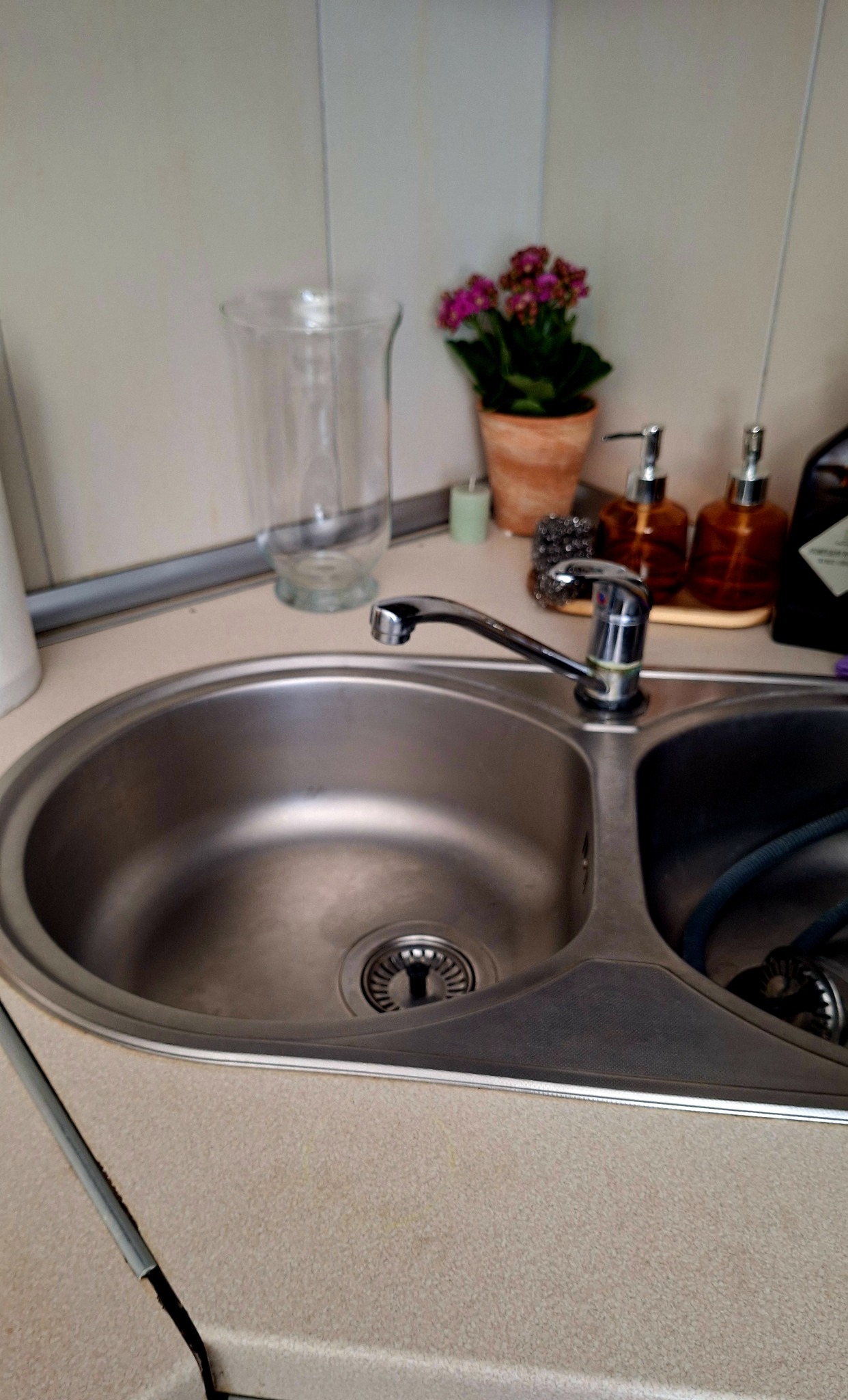 Błyszczący, dwukomorowy zlewozmywak ze stali nierdzewnej, bateria, kwiat w doniczce i dozowniki mydła na blacie w kuchni. Czystość i porządek.