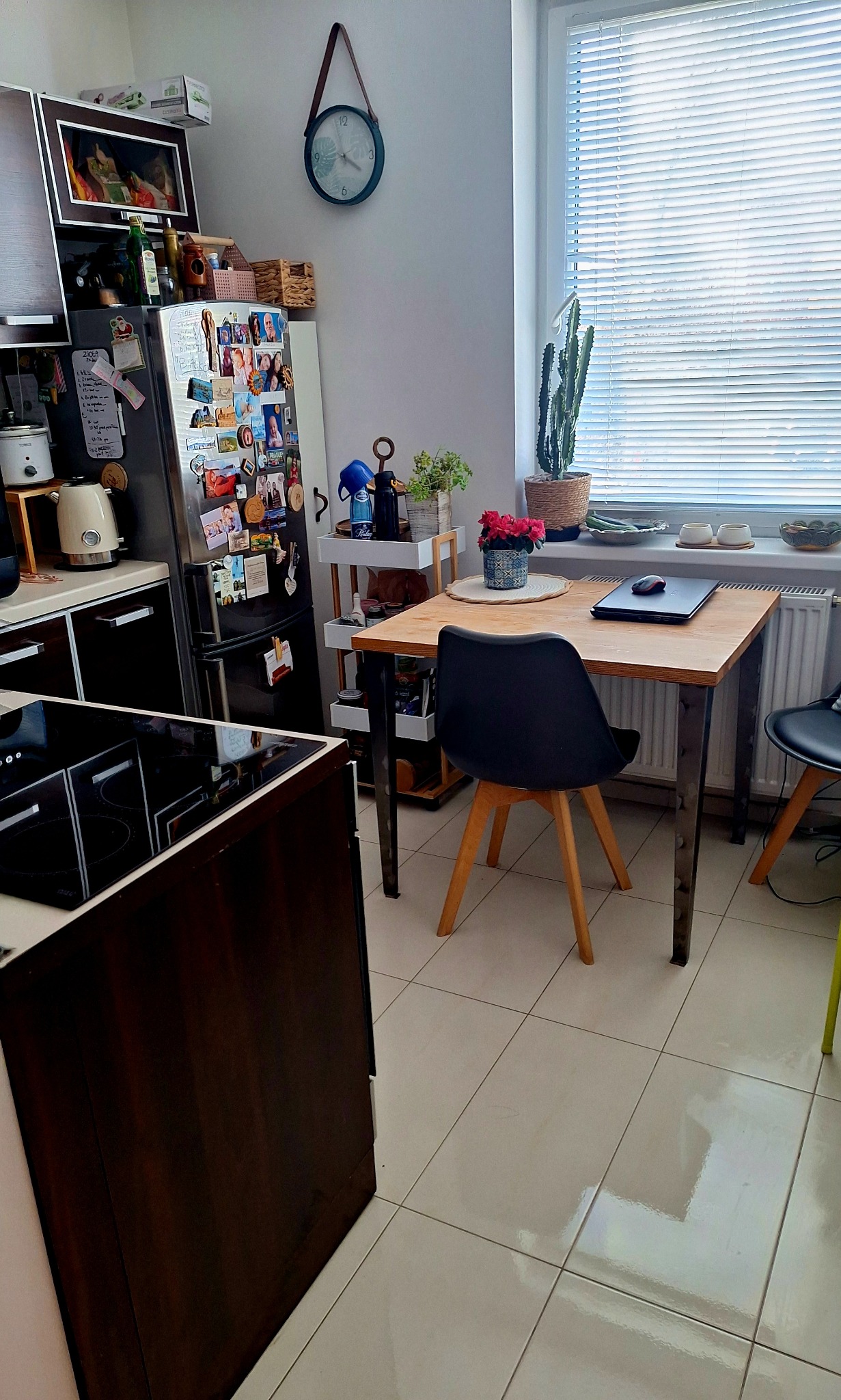 Ustawna kuchnia z drewnianym stołem, czarnym krzesłem i kwiatami w doniczce. Na blacie laptop. W tle okno z żaluzjami i lodówka z magnesami.