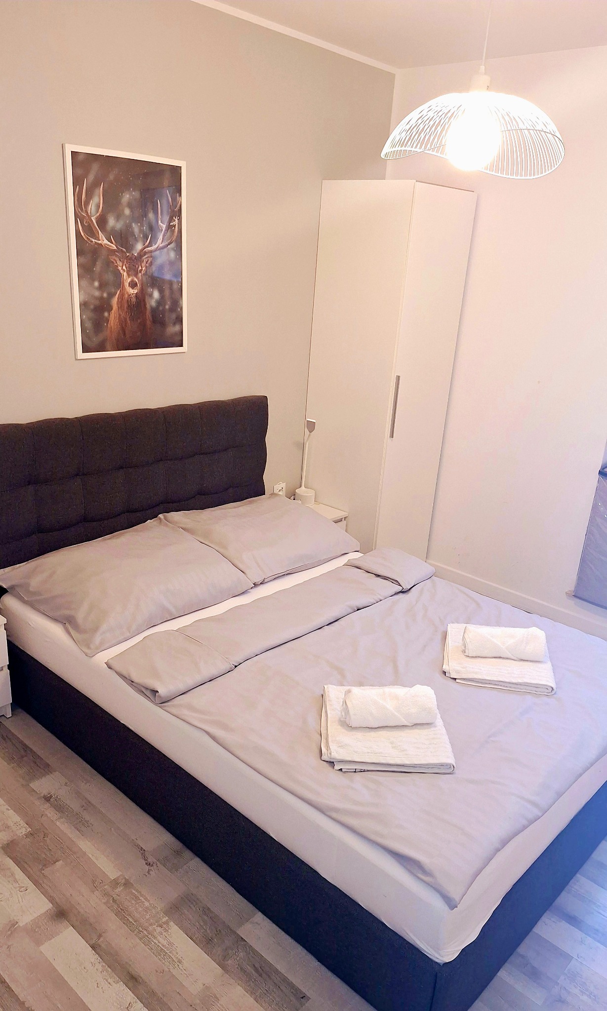 Jasna sypialnia z łóżkiem, szarą pościelą i ręcznikami. Na ścianie plakat z jeleniem, obok biała szafa. Minimalistyczny design i czystość.