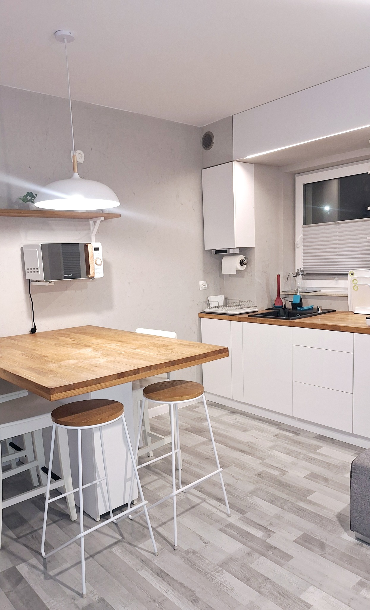 Jasna kuchnia z drewnianym blatem, białymi szafkami i hokerami. Na ścianie mikrofala i półka. Minimalistyczny design, szare panele podłogowe.