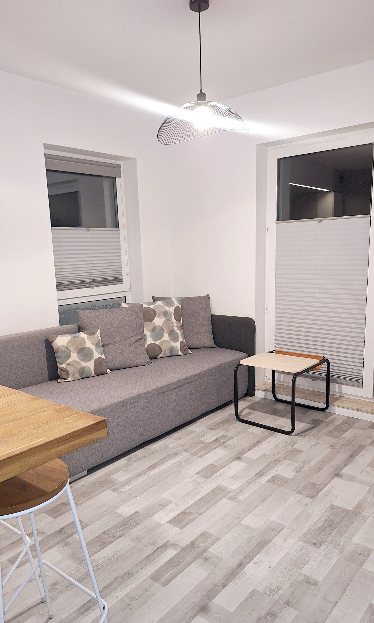 Jasne wnętrze pokoju dziennego z szarą sofą, drewnianym stolikiem i krzesłem barowym. Na podłodze panele imitujące drewno. Okna z żaluzjami plisowanymi.