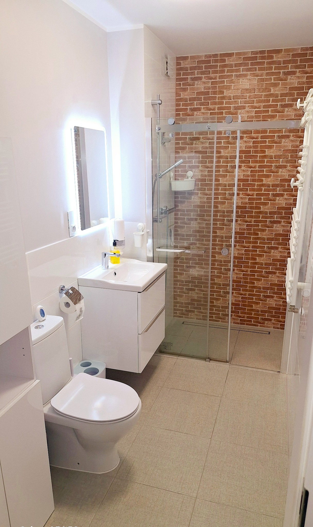 Jasna łazienka z kabiną prysznicową wykończoną cegłą, białą armaturą i podświetlanym lustrem. Nowoczesny design z akcentem rustykalnym.