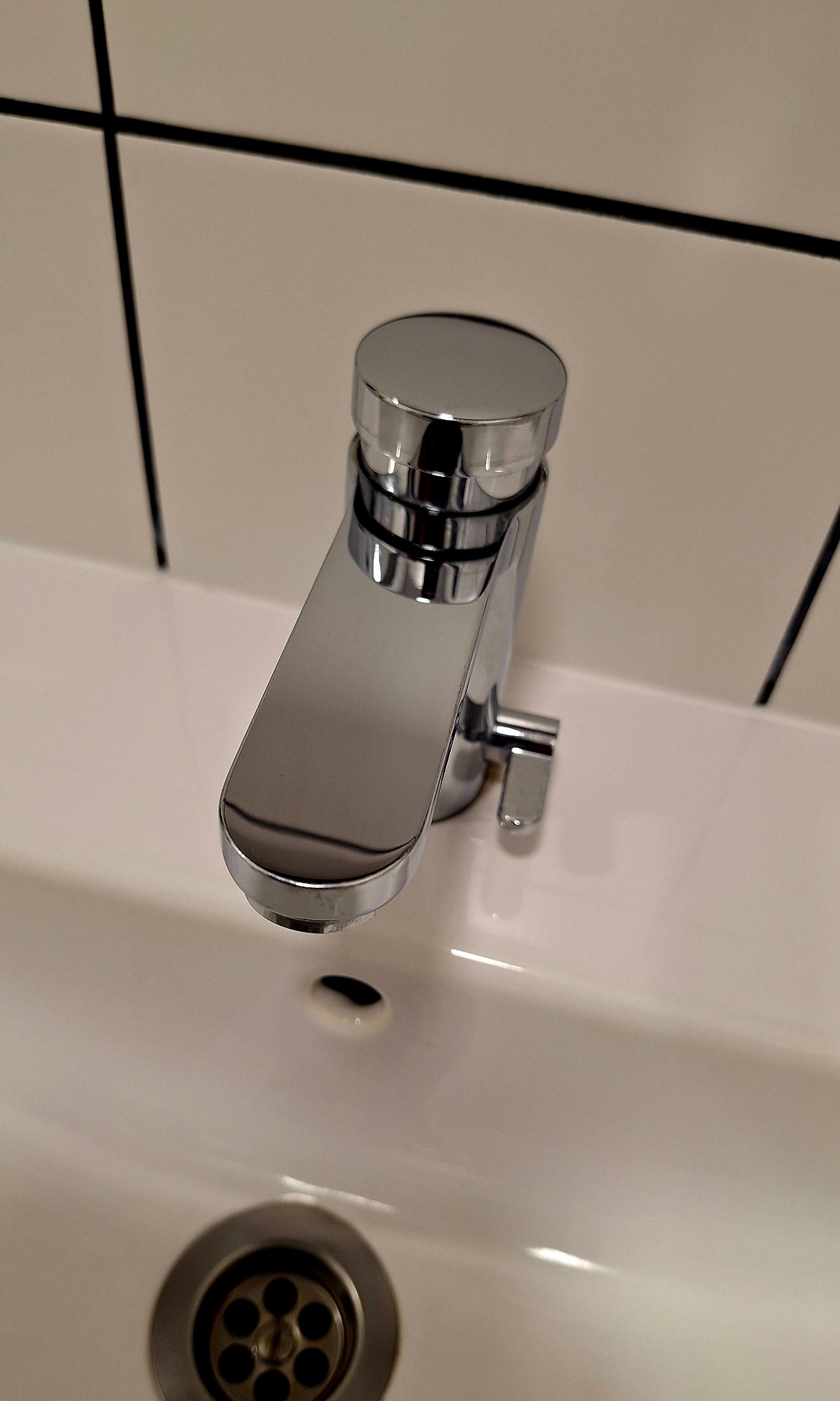 Błyszcząca, chromowana bateria umywalkowa z odpływem na białej umywalce, na tle jasnych płytek z ciemnymi fugami. Minimalistyczny design, czystość i porządek.