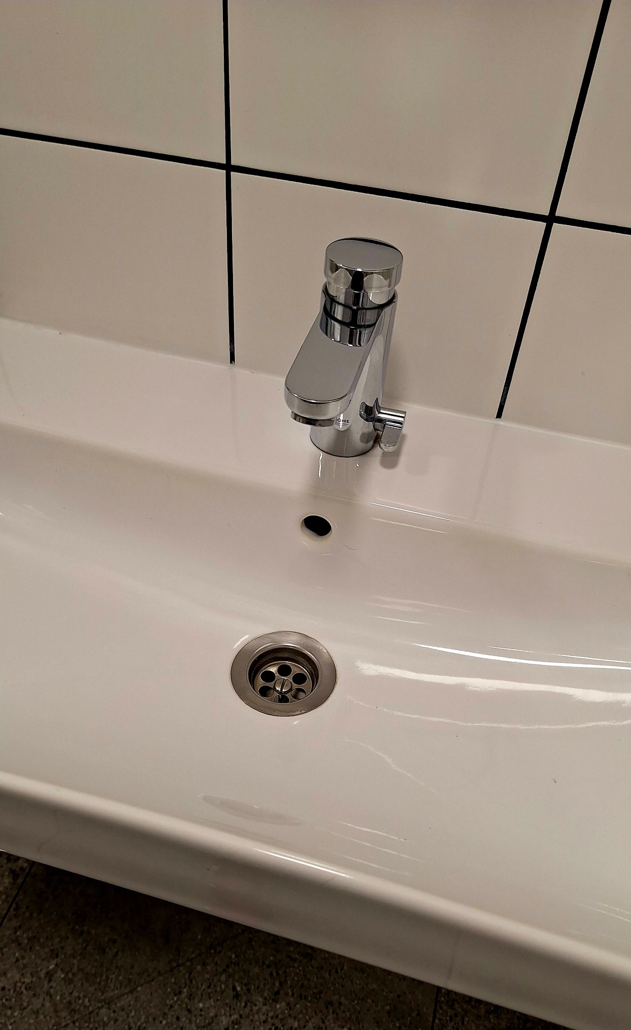 Biała umywalka z chromowaną baterią Grohe, widok z góry. Minimalistyczny design, czyste linie, płytki w tle. Odpływ z metalową kratką.