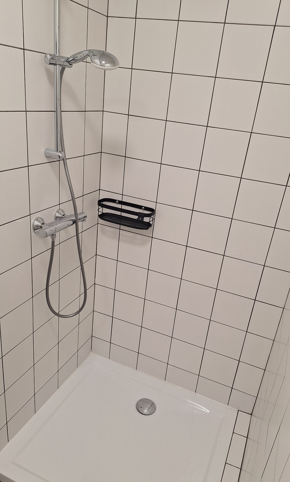 Biały brodzik z odpływem, chromowana bateria prysznicowa i czarna półka na kosmetyki na tle białych, kwadratowych płytek z ciemną fugą w kabinie prysznicowej.