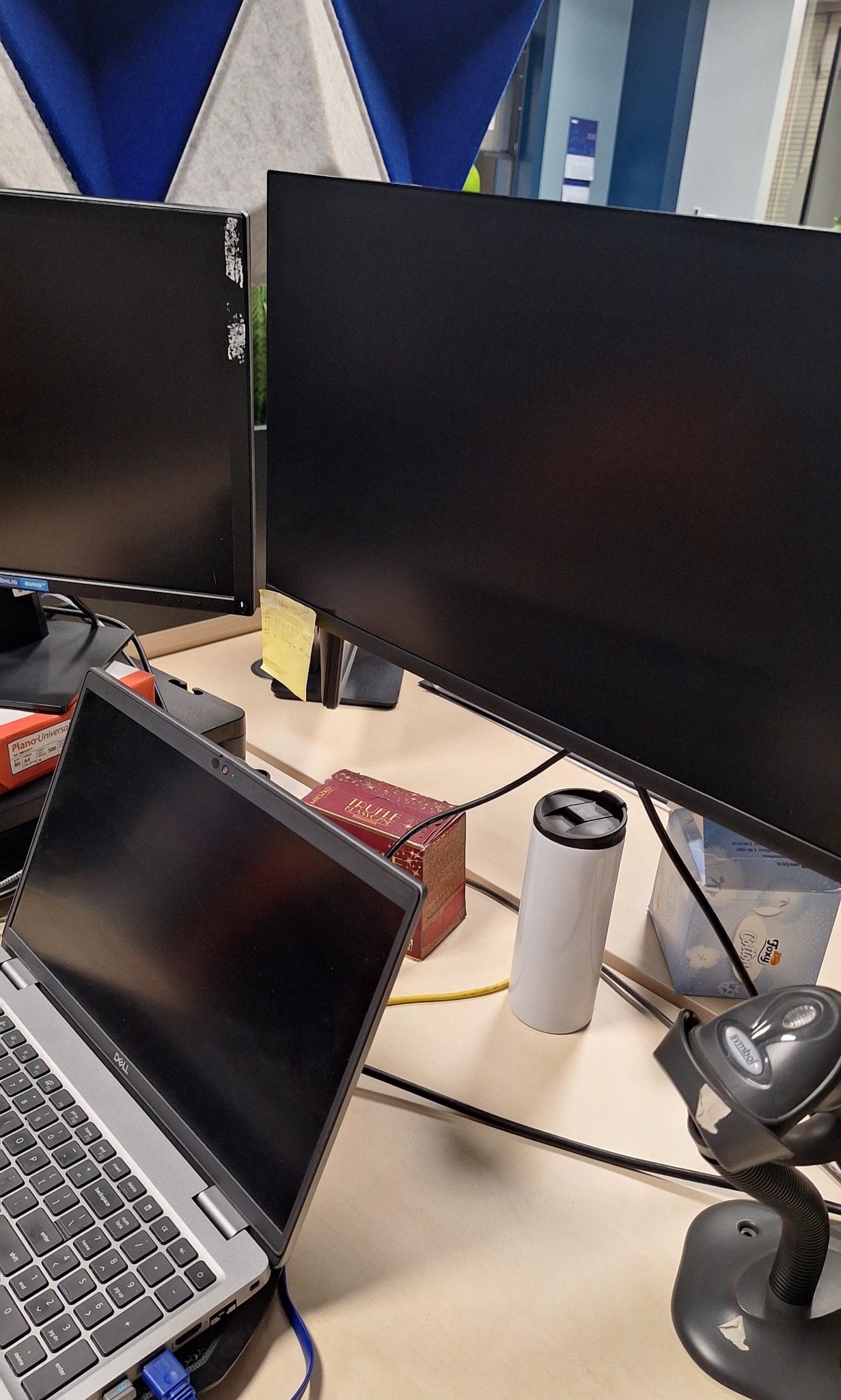 Biurko z dwoma monitorami, laptopem Dell, skanerem kodów kreskowych, pudełkiem Trufle i kubkiem termicznym. Jeden z monitorów ma ślady po naklejkach. Tło stanowią panele akustyczne.