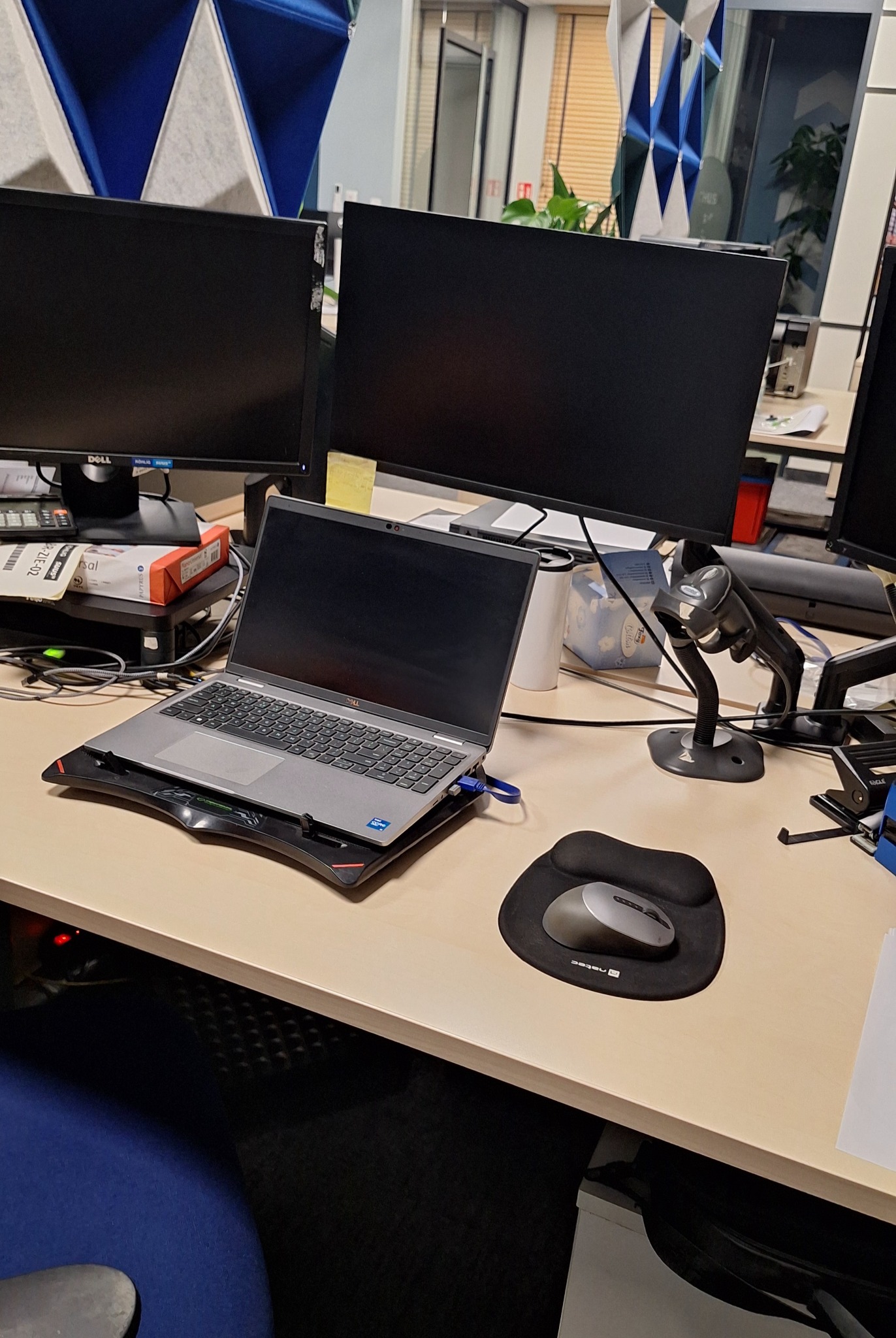 Stanowisko pracy biurowej z laptopem na podstawce, dwoma monitorami Dell, skanerem kodów kreskowych i podkładką pod mysz. Widoczne biurko i krzesło.