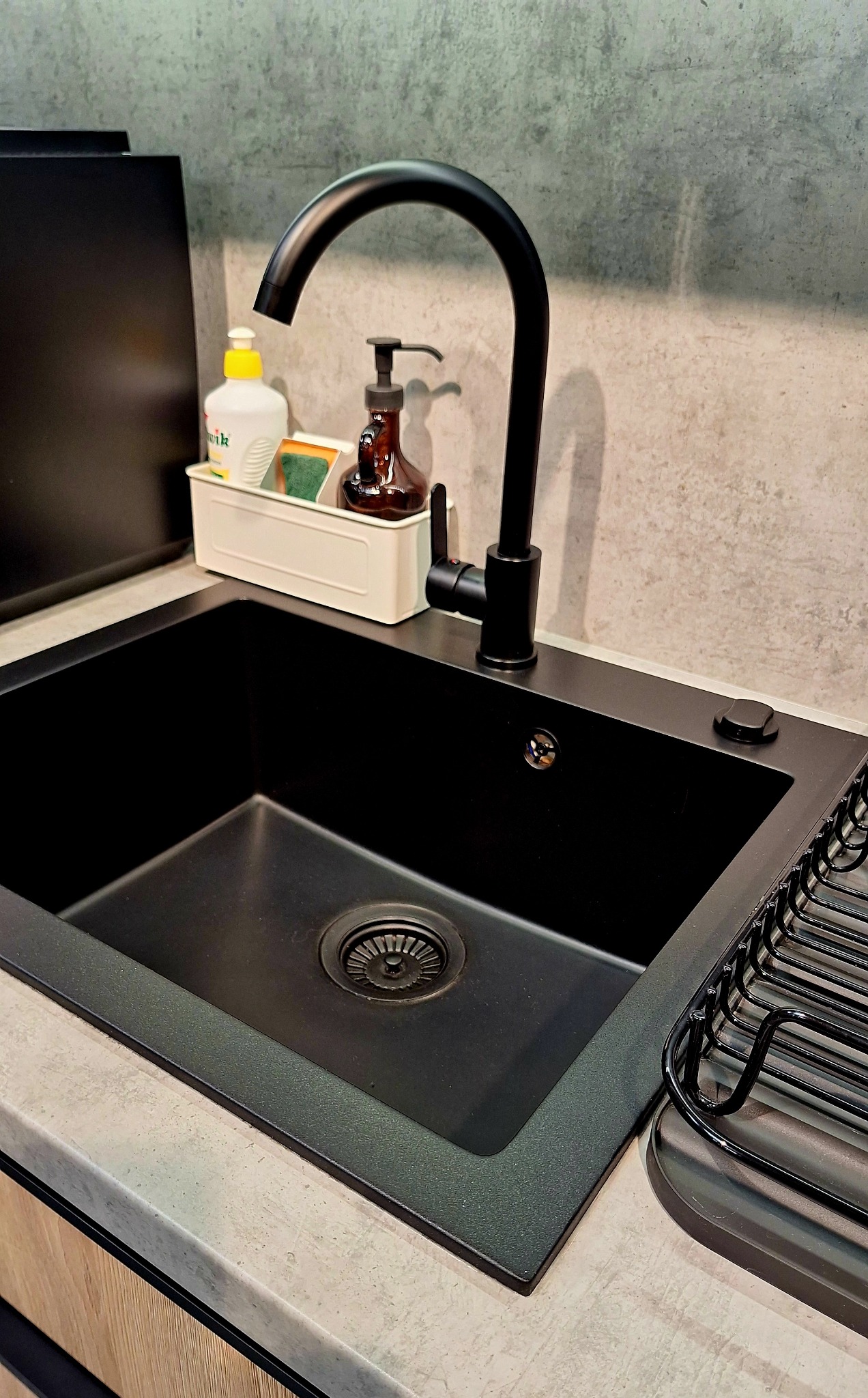 Czarny, minimalistyczny zlew granitowy z baterią i ociekaczem. Na blacie dozownik mydła i płyn do naczyń. Nowoczesna kuchnia z szarym blatem i betonową ścianą w tle.