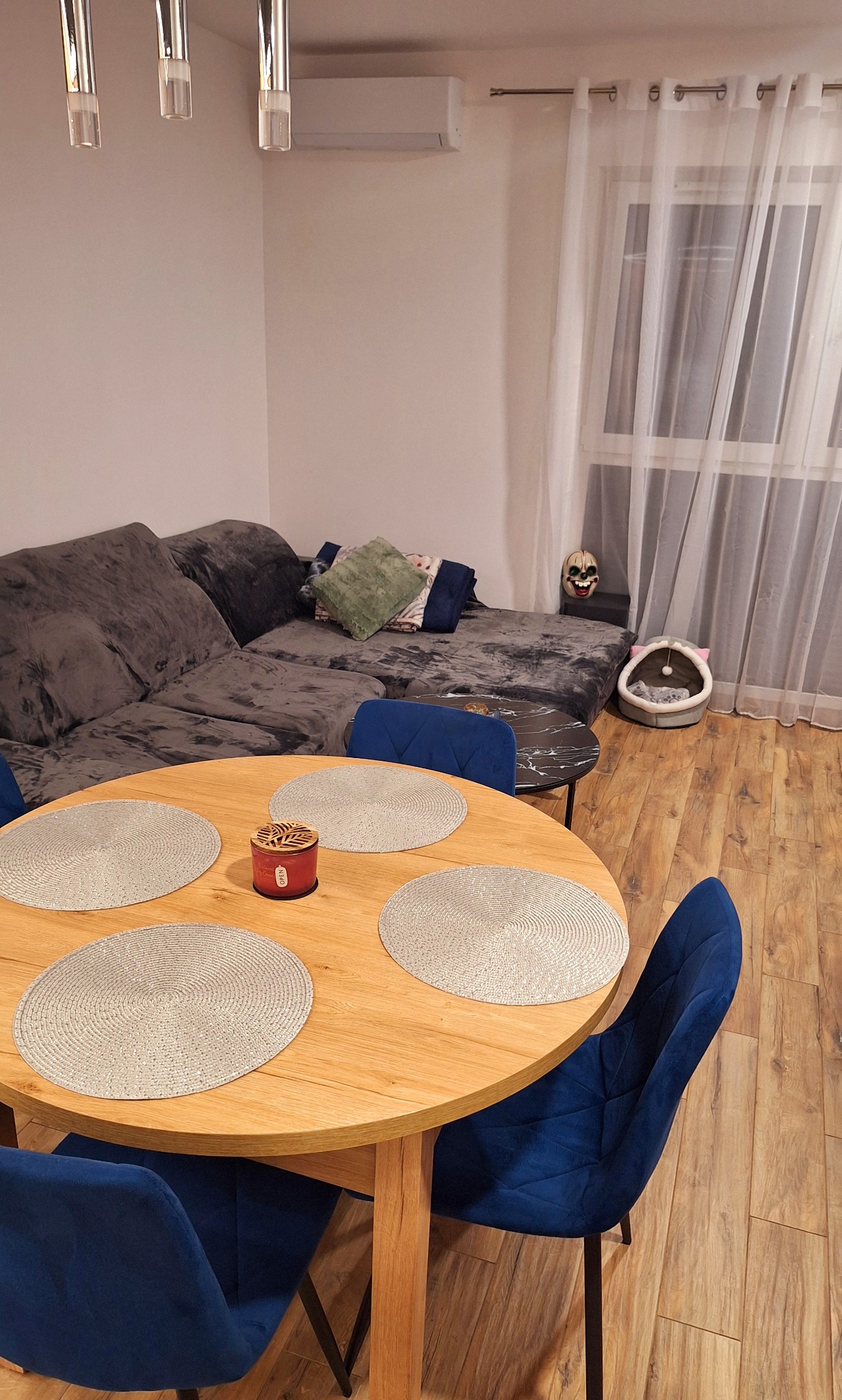 Jasne wnętrze pokoju z okrągłym stołem, niebieskimi krzesłami i szarą sofą. Na stole podkładki i świeca. W tle okno z firanką i legowisko dla kota.