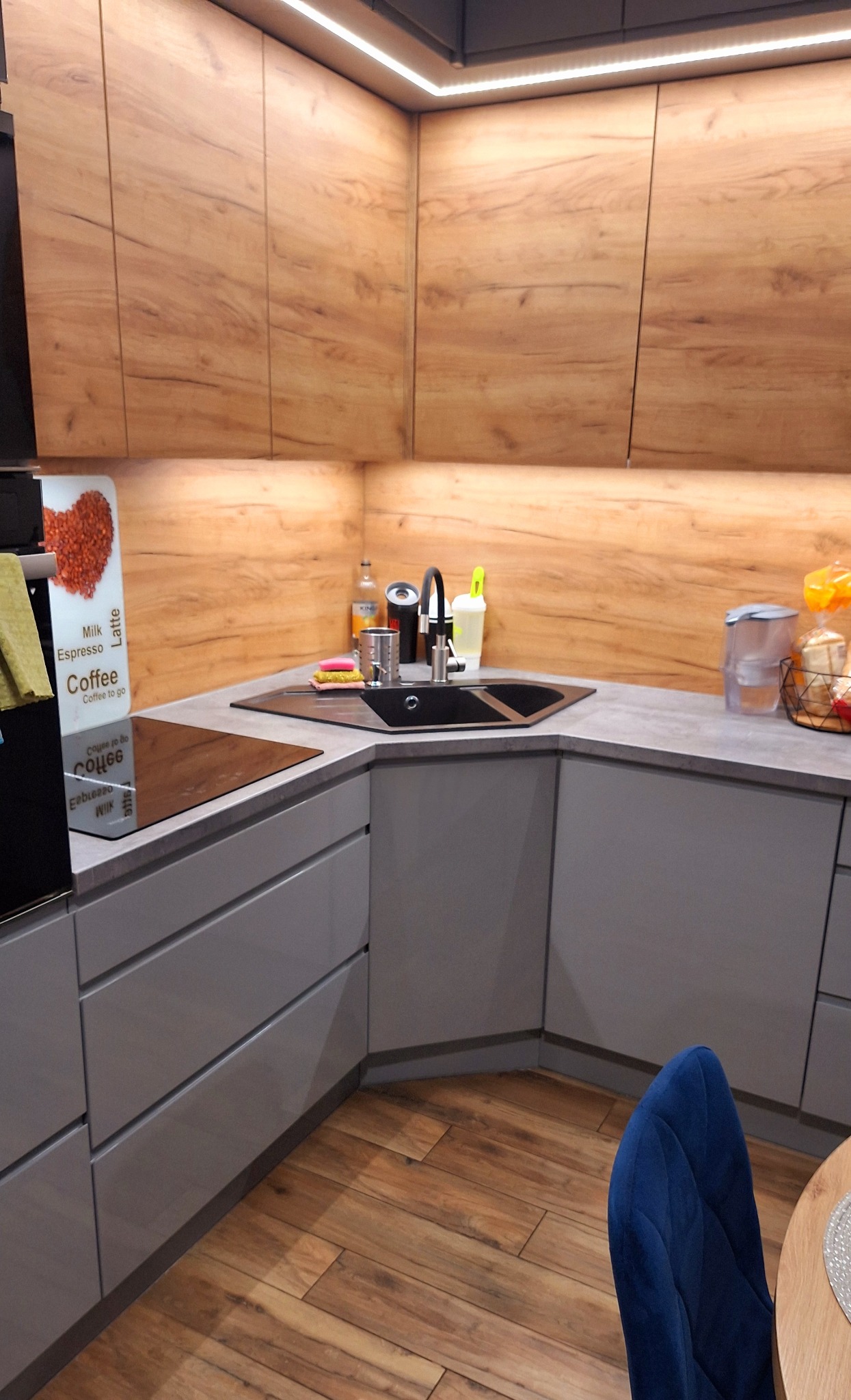 Nowoczesna kuchnia w odcieniach szarości i drewna. Uwagę przykuwa czarny zlew narożny i oświetlenie LED pod szafkami, tworzące ciepłą atmosferę. Widoczny fragment krzesła.