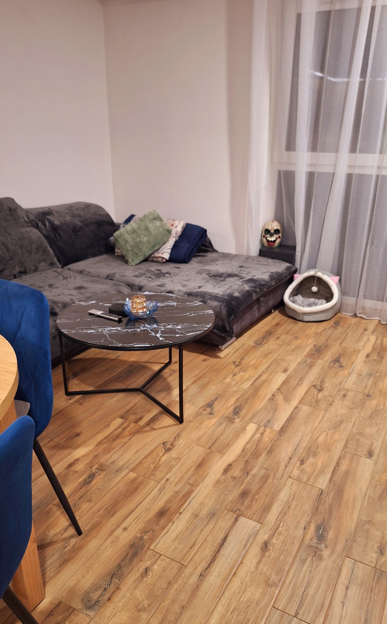 Uporządkowany salon z szarą sofą, okrągłym stolikiem z marmurowym blatem, drewnianą podłogą i dekoracjami, w tym maską czaszki przy oknie.