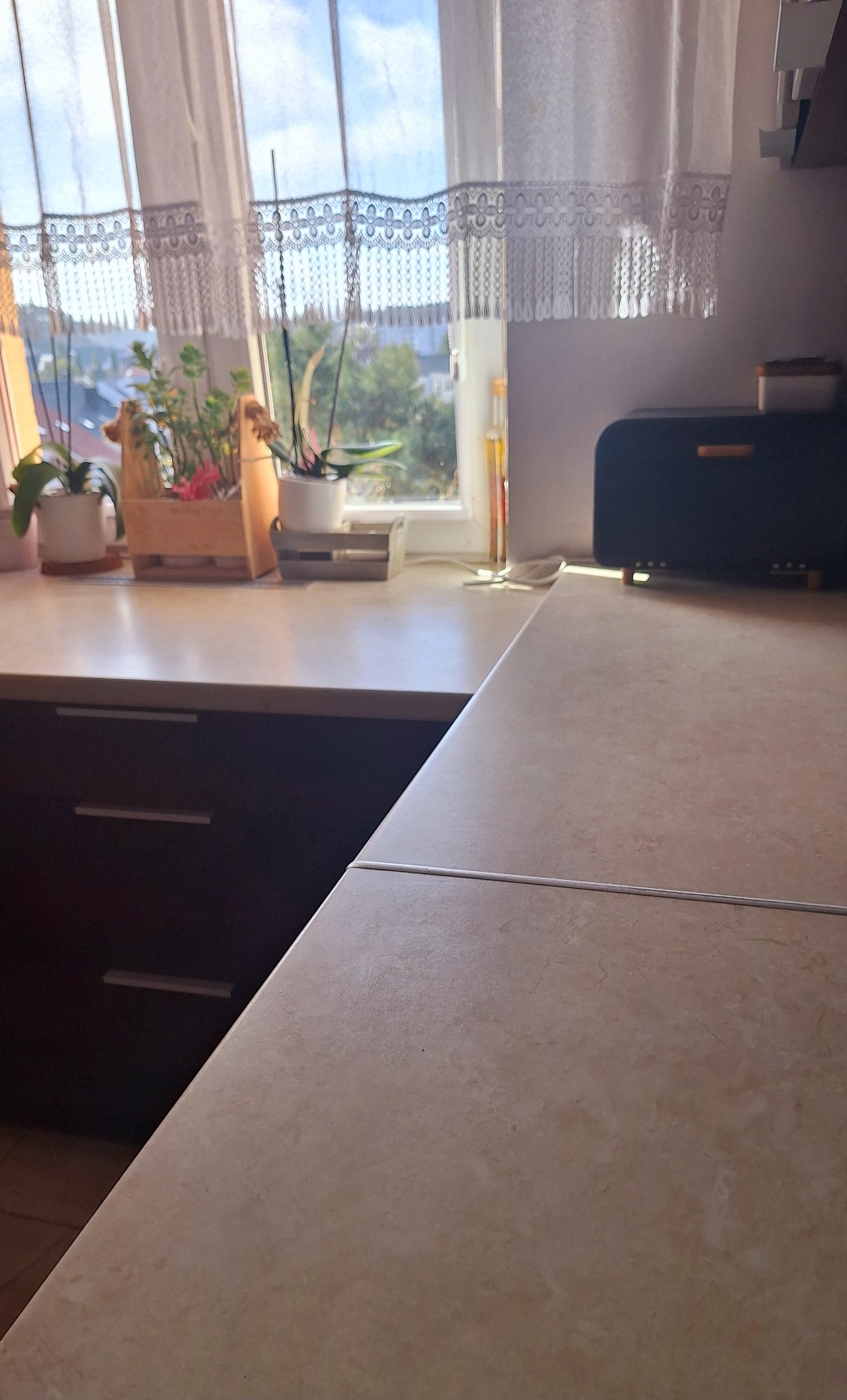 Jasna kuchnia z roślinami w oknie i drewnianym chlebakiem. Blat z delikatnym wzorem, ciemne szafki. Dzień, naturalne światło, firanka z koronką.