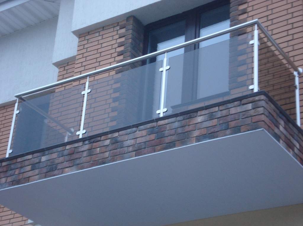Szklana balustrada balkonowa z metalowymi słupkami i poręczą, zamontowana na ceglanym balkonie z szarą podłogą. Widok z dołu na fragment elewacji budynku.