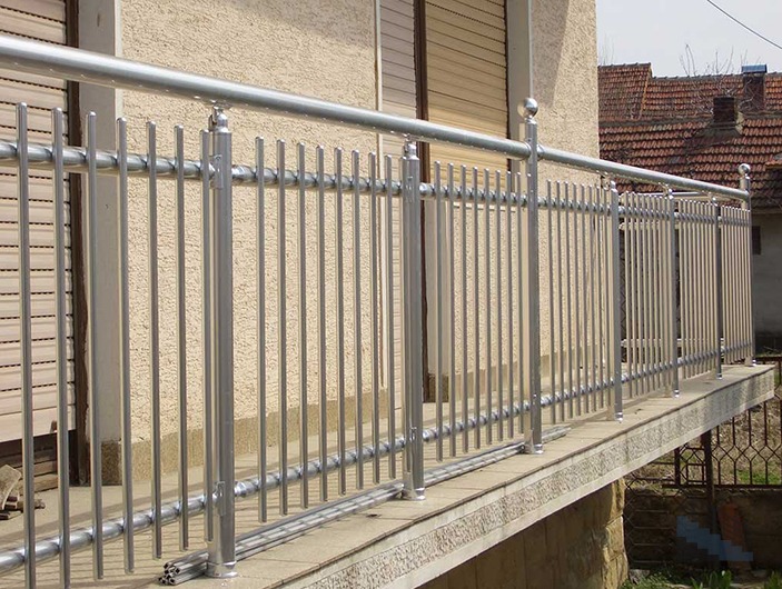 Balkon z nowoczesną, srebrną balustradą z pionowych prętów i kulistych zakończeń, na tle beżowej elewacji budynku z zamkniętymi roletami.