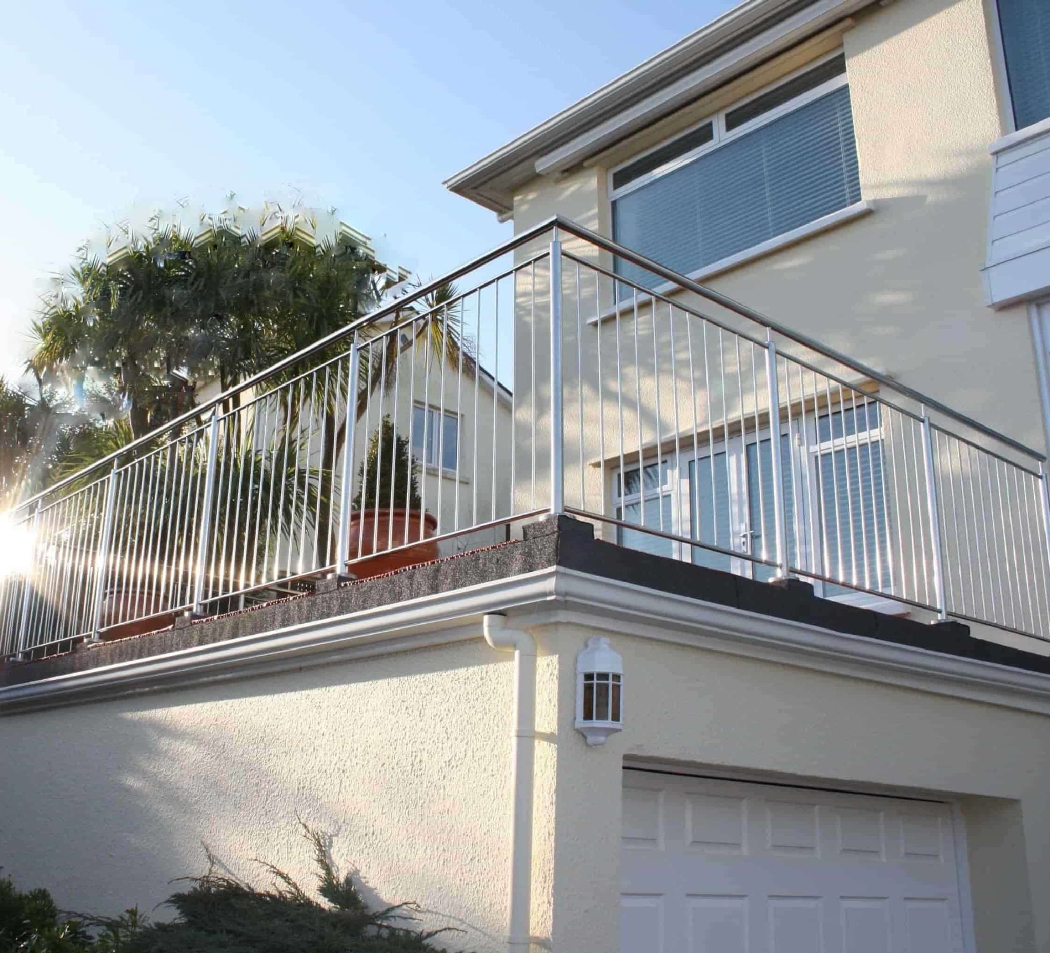 Balkon z nowoczesną, nierdzewną balustradą. Słoneczne odbicie na metalowych prętach. Widok z dołu na jasny budynek z garażem i roślinnością w tle.